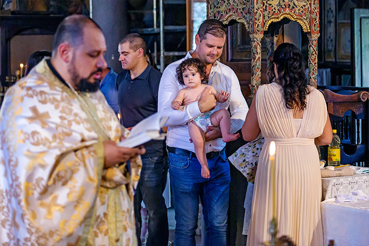 Отецът чете от требника, докато събличат момиченцето за нейното кръщение в купела.